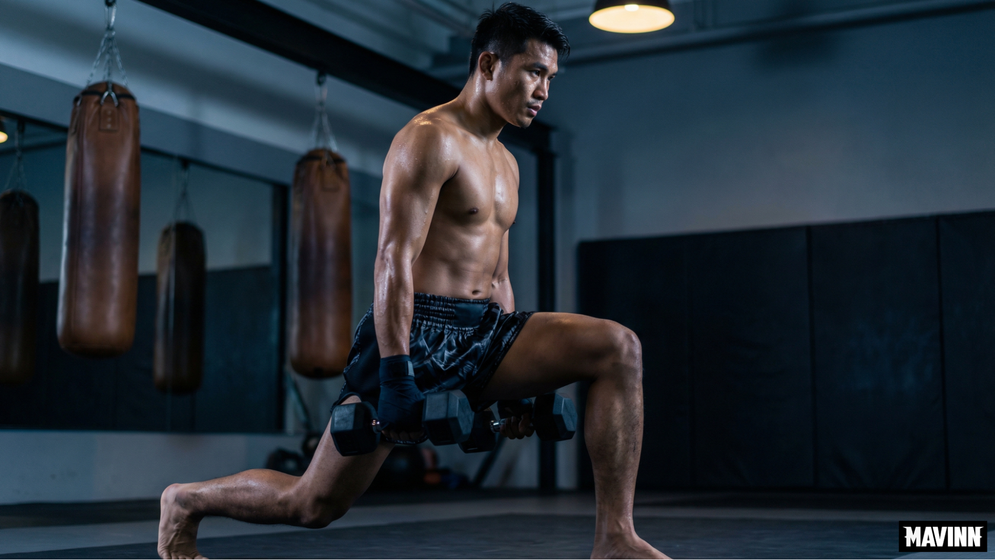 Shirtless athlete performing dumbbell lunges in a gym during a muay thai workout